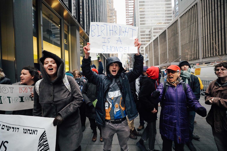 A young man wearing a hooded black jacket holds up a sign saying "BIRTHRIGHT IS A SHONDE" in a crowd of protesters in midtown Manhattan.